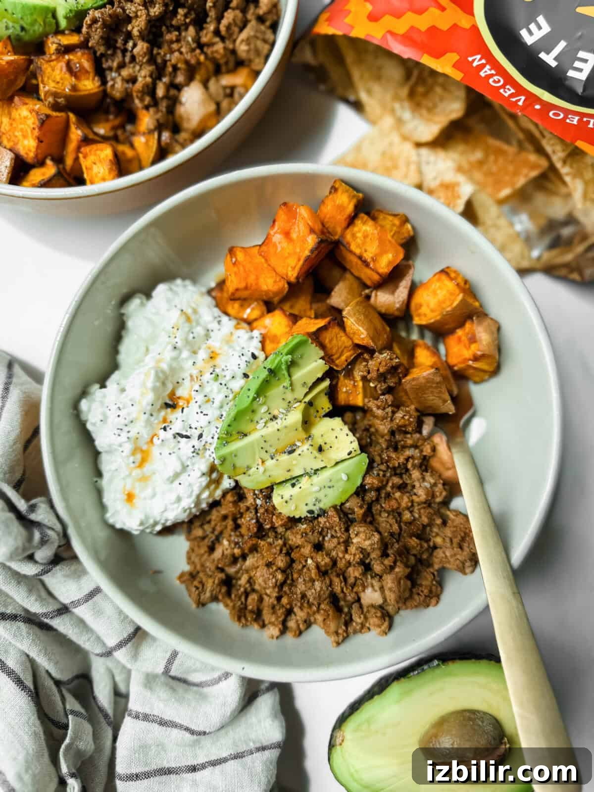A bowl with ground beef, roasted sweet potatoes, cottage cheese, and avocado slices sits next to an open bag of tortilla chips and a halved avocado.