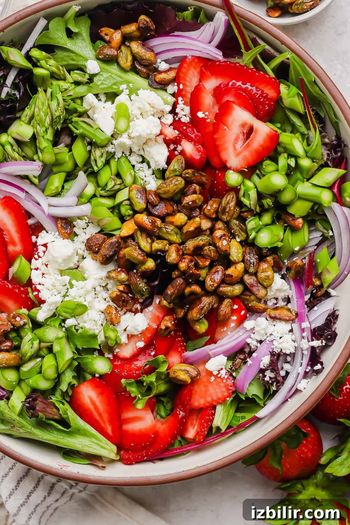 Crisp Summer Salad with Honey-Roasted Pistachios 2 A salad with mixed greens, sliced strawberries, red onions, asparagus, edamame, crumbled cheese, and roasted pistachios in a bowl.