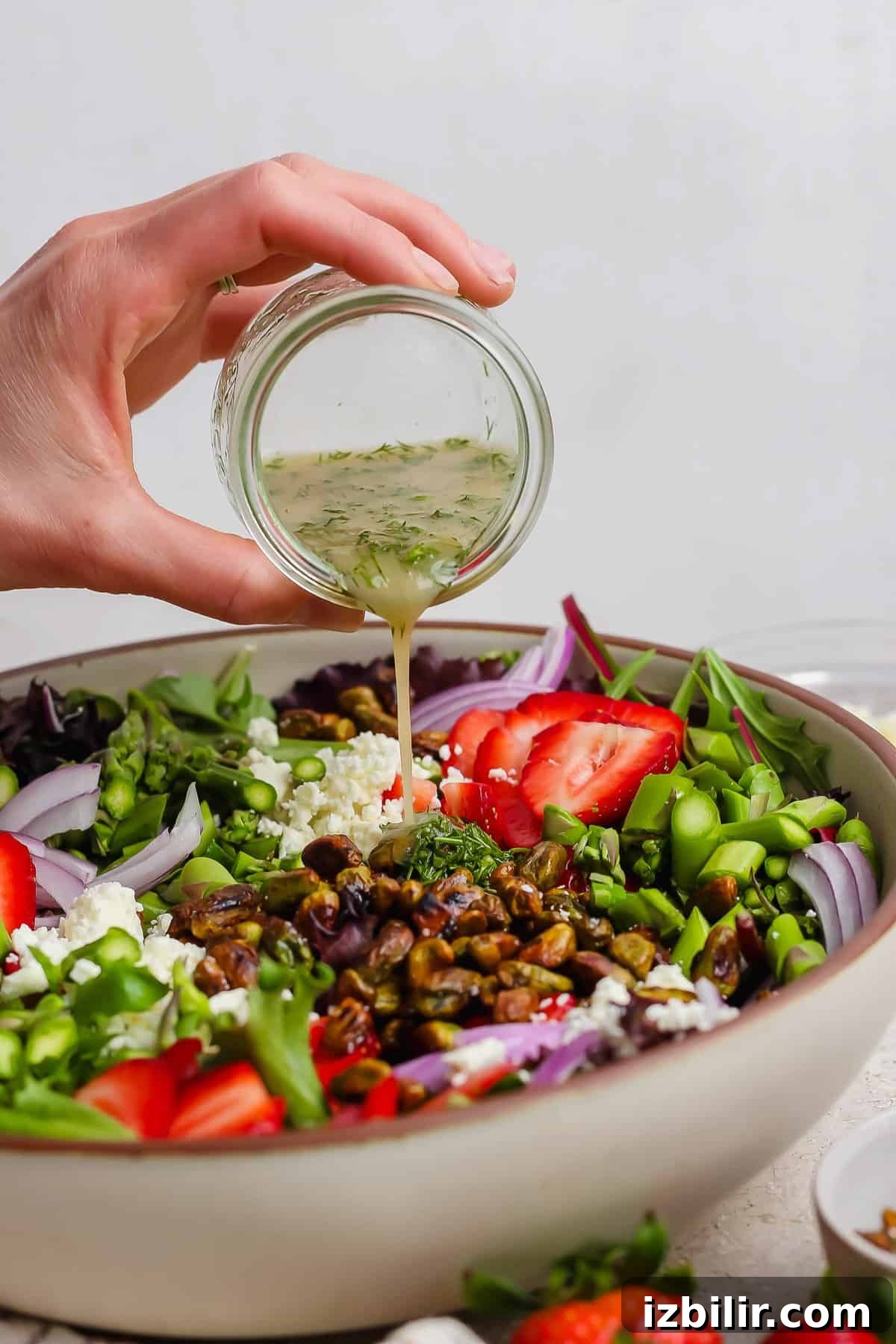 Crisp Summer Salad with Honey-Roasted Pistachios 4 A hand pours vinaigrette from a small jar onto a salad with strawberries, feta cheese, red onions, pistachios, and greens in a large bowl.