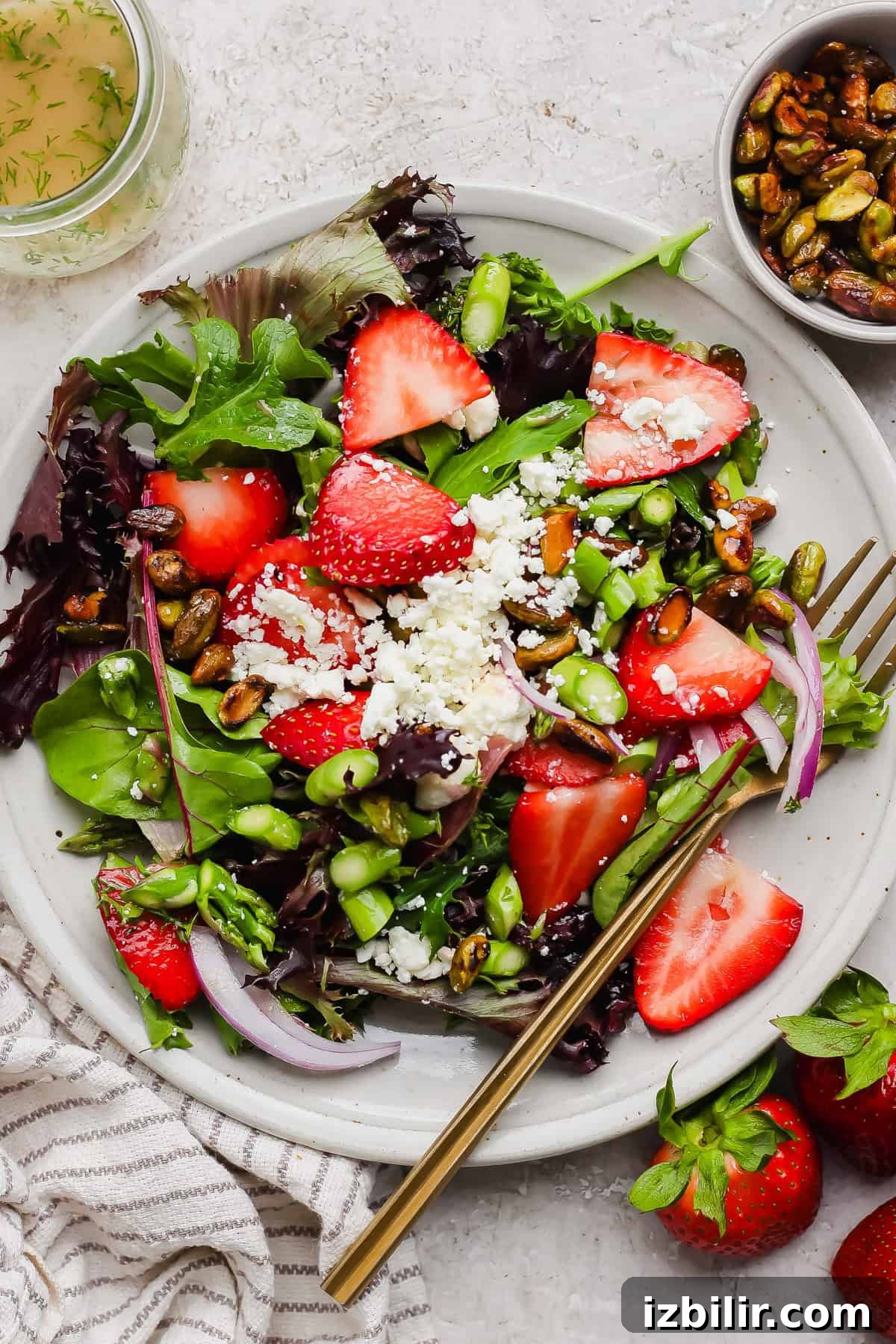 Crisp Summer Salad with Honey-Roasted Pistachios 5 A plate of mixed greens salad with sliced strawberries, crumbled cheese, pistachios, red onion, and a fork, next to a bowl of nuts and fresh strawberries.