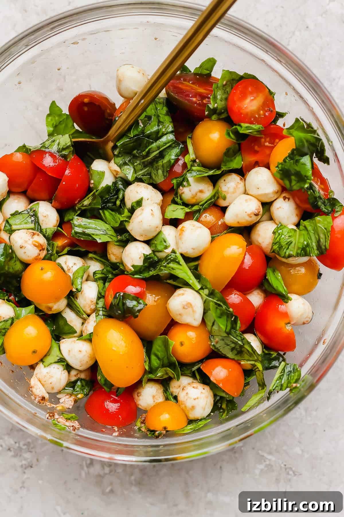 Seared Balsamic Steak Caprese with Fresh Mozzarella 3 A clear glass bowl filled with cherry tomatoes, mozzarella balls, fresh basil leaves, and a spoon, on a light surface.
