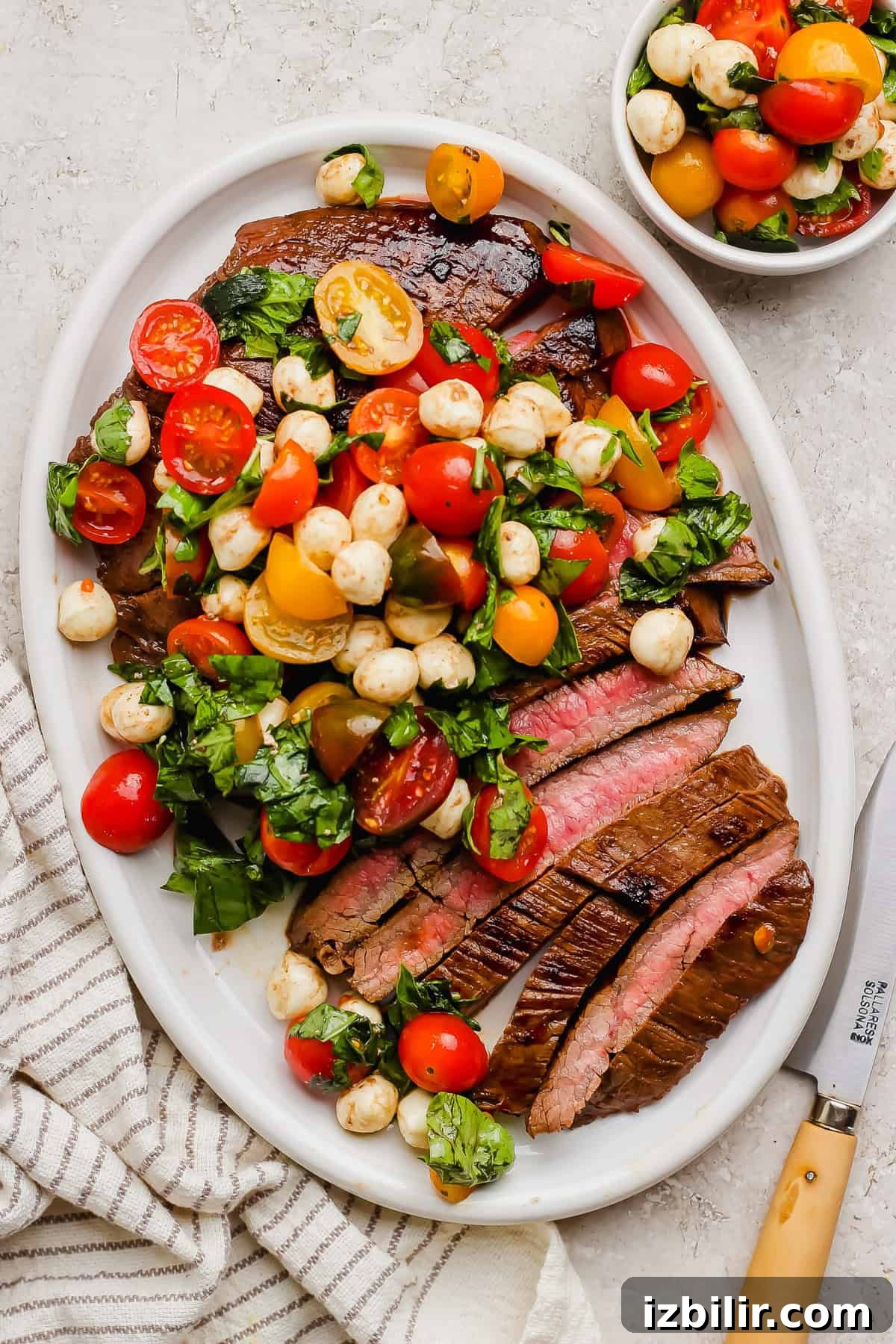 Seared Balsamic Steak Caprese with Fresh Mozzarella 6 Sliced medium-rare steak on a white oval platter, topped with a tomato, mozzarella, and basil salad; a matching small bowl is beside it. A striped towel and knife are nearby.