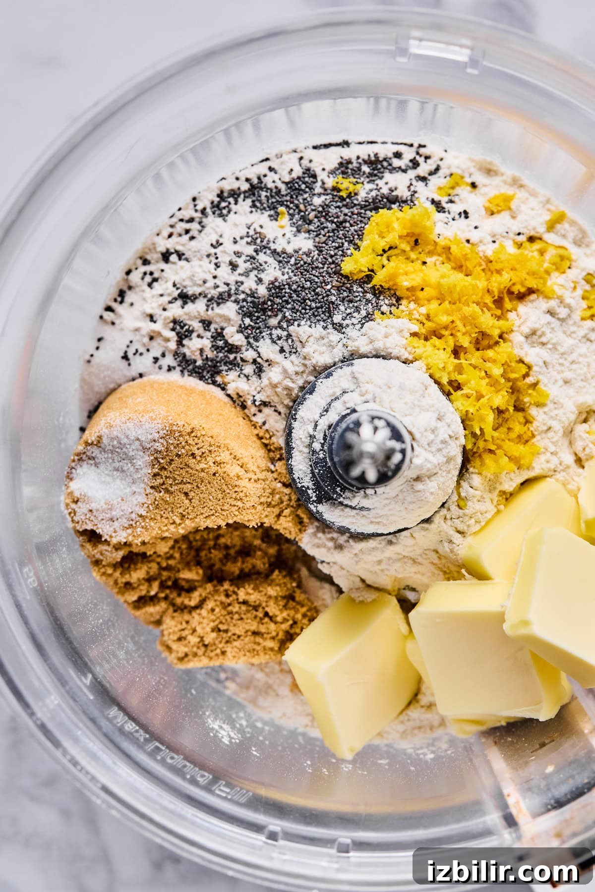 A food processor bowl containing flour, brown sugar, poppy seeds, lemon zest, salt, and cubes of butter, ready to be mixed.