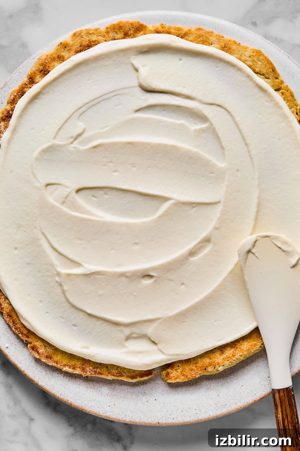 A partially frosted round cake on a plate, with a spatula spreading white frosting over the surface.