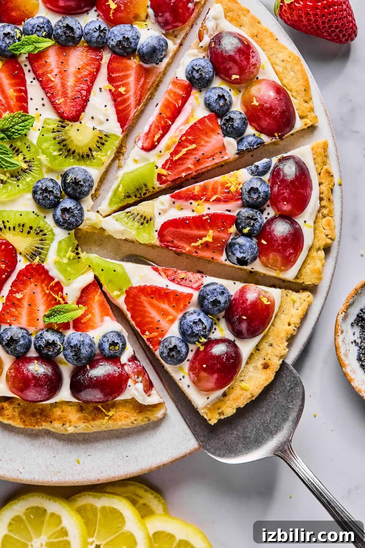 A fruit pizza topped with sliced strawberries, grapes, kiwi, and blueberries on a cream base, with a pie server lifting one slice from the plate.