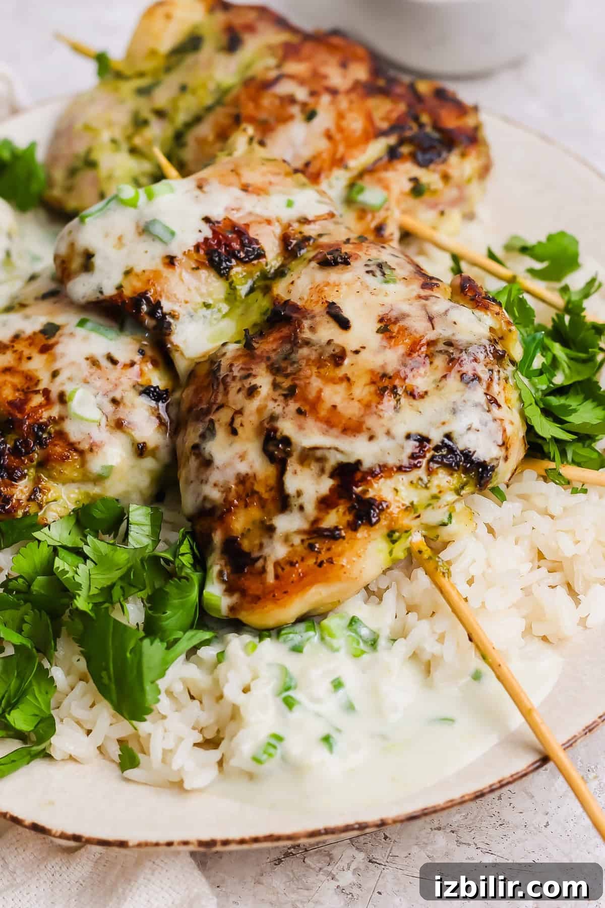 Three grilled chicken skewers topped with a creamy white sauce served over white rice and garnished with fresh cilantro.