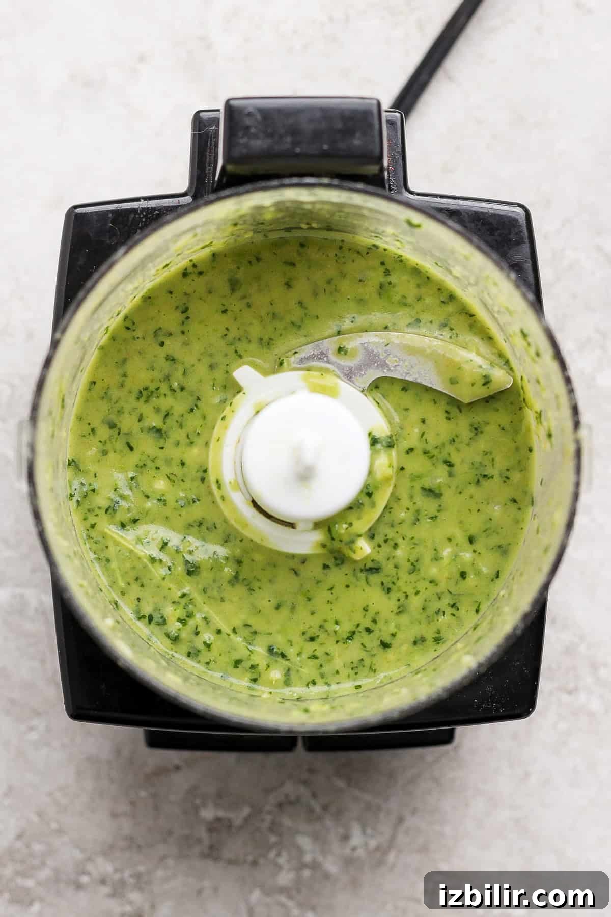 Food processor containing a green sauce or mixture with visible herbs on a light countertop.