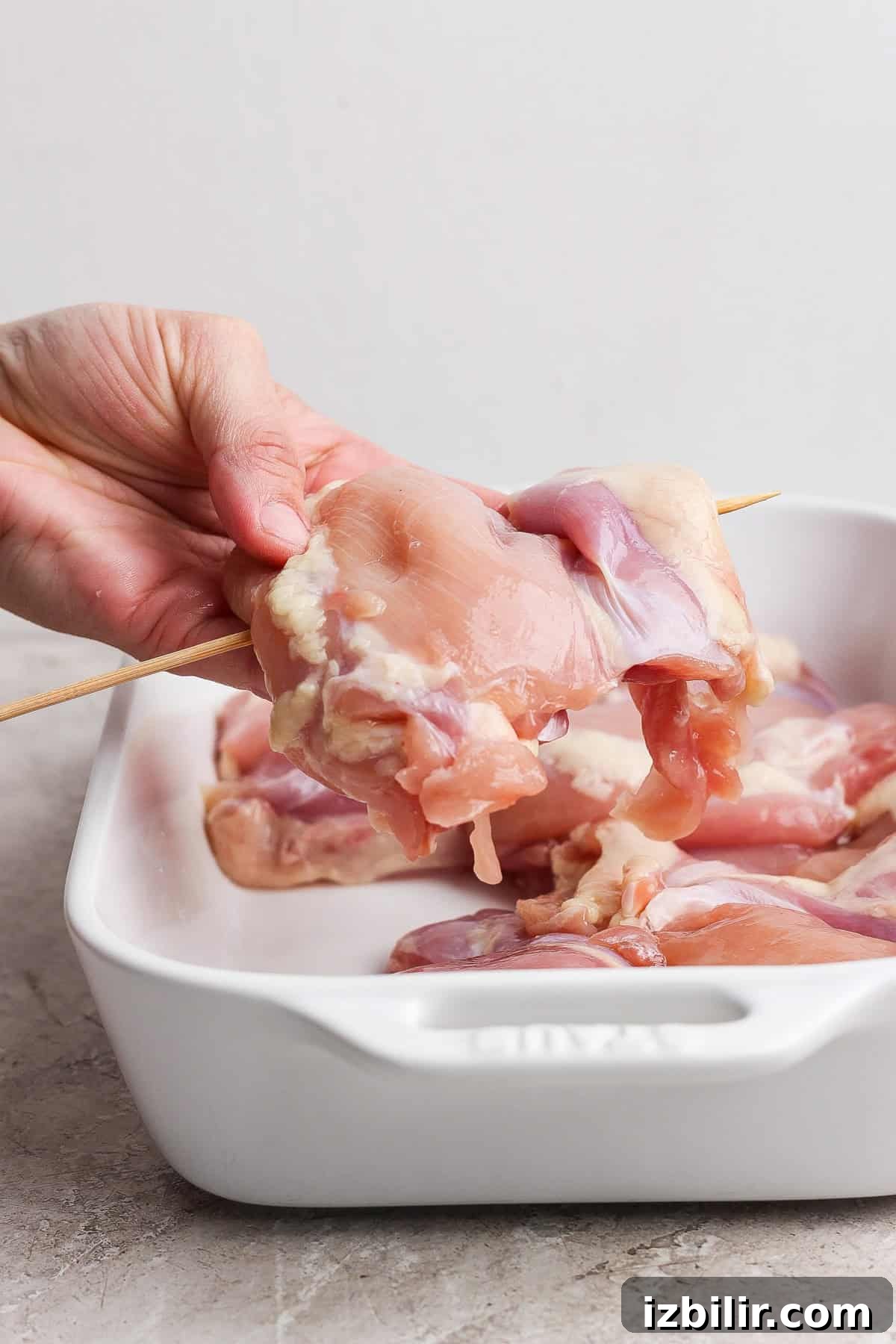 Hand holding raw chicken piece on a wooden skewer over a baking dish with more raw chicken pieces.