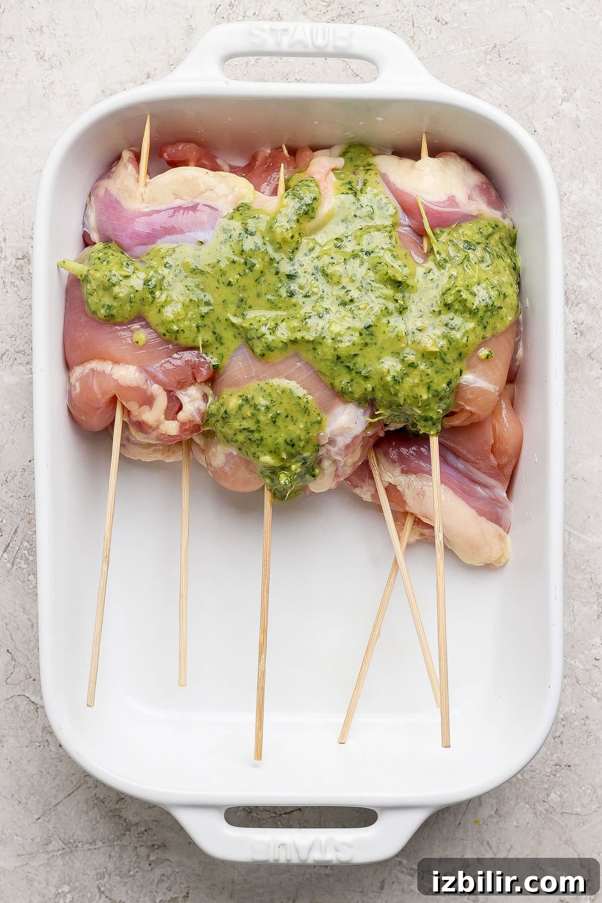 Raw chicken thighs skewered on wooden sticks in a white baking dish, topped with a green herb sauce.