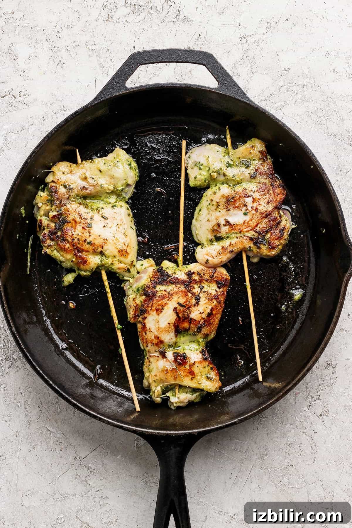 Three grilled, stuffed chicken thighs on wooden skewers cooking in a cast iron skillet.