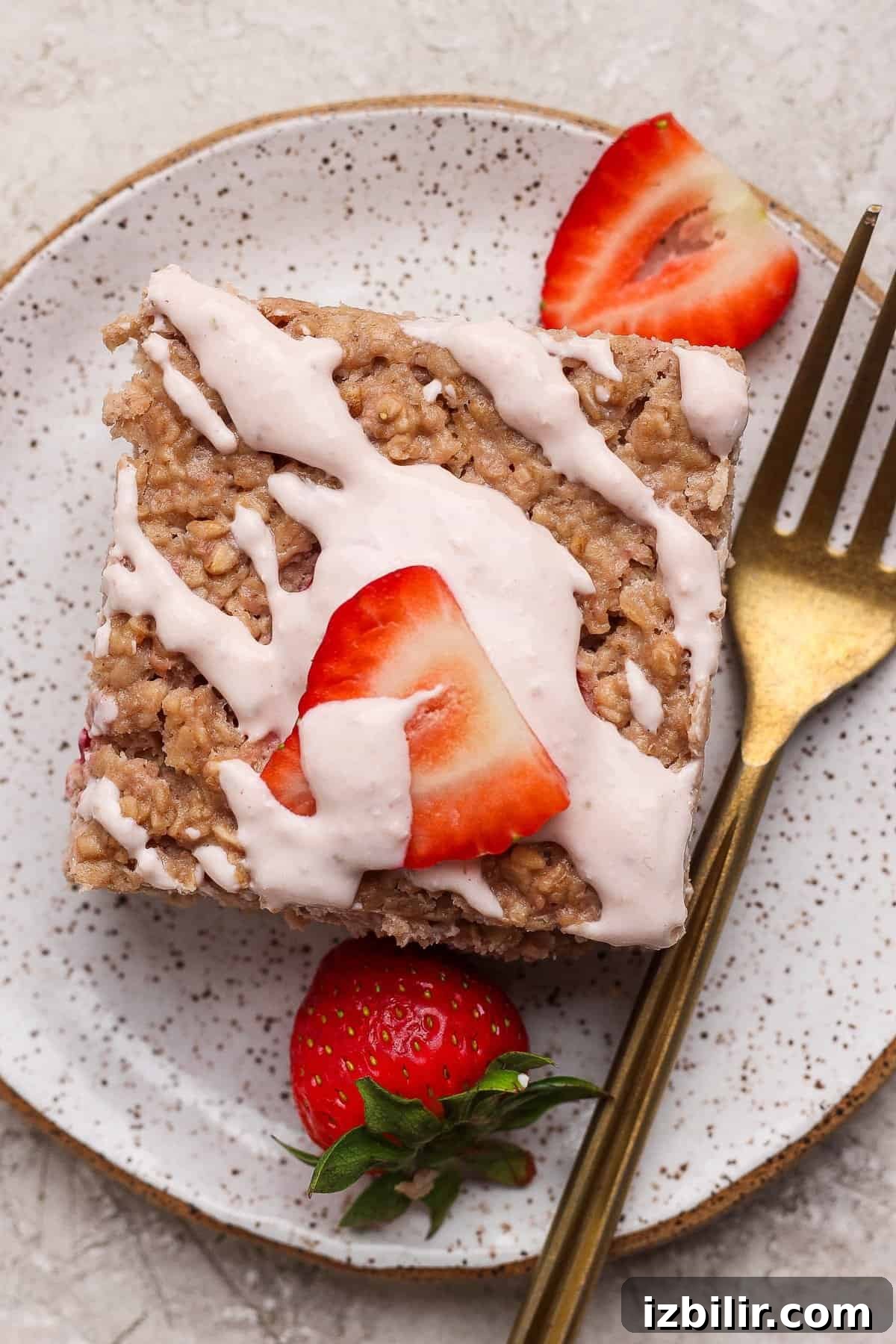 Strawberry Cheesecake Baked Oats Recipe with Creamy Filling 2 A square piece of oatmeal bake topped with white icing and sliced strawberries on a speckled plate, with a gold fork beside it.