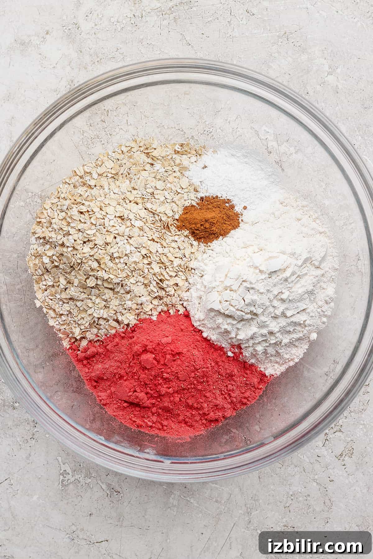 Strawberry Cheesecake Baked Oats Recipe with Creamy Filling 3 A glass bowl containing oats, white flour, pink powder, cinnamon, baking powder, and baking soda on a light surface.