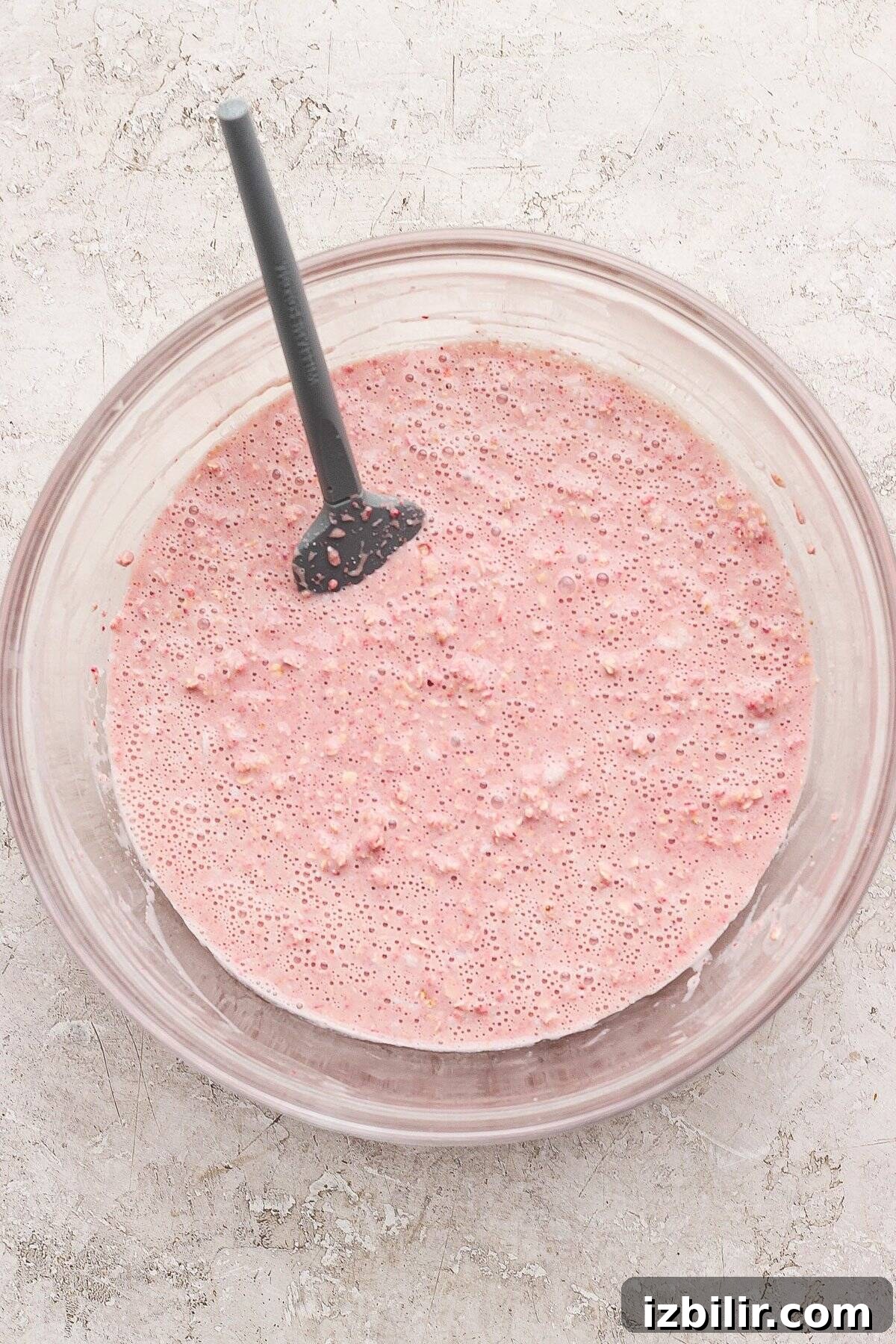 Strawberry Cheesecake Baked Oats Recipe with Creamy Filling 4 A glass bowl filled with a pink, bubbly batter mixture and a black spatula, placed on a light textured surface.