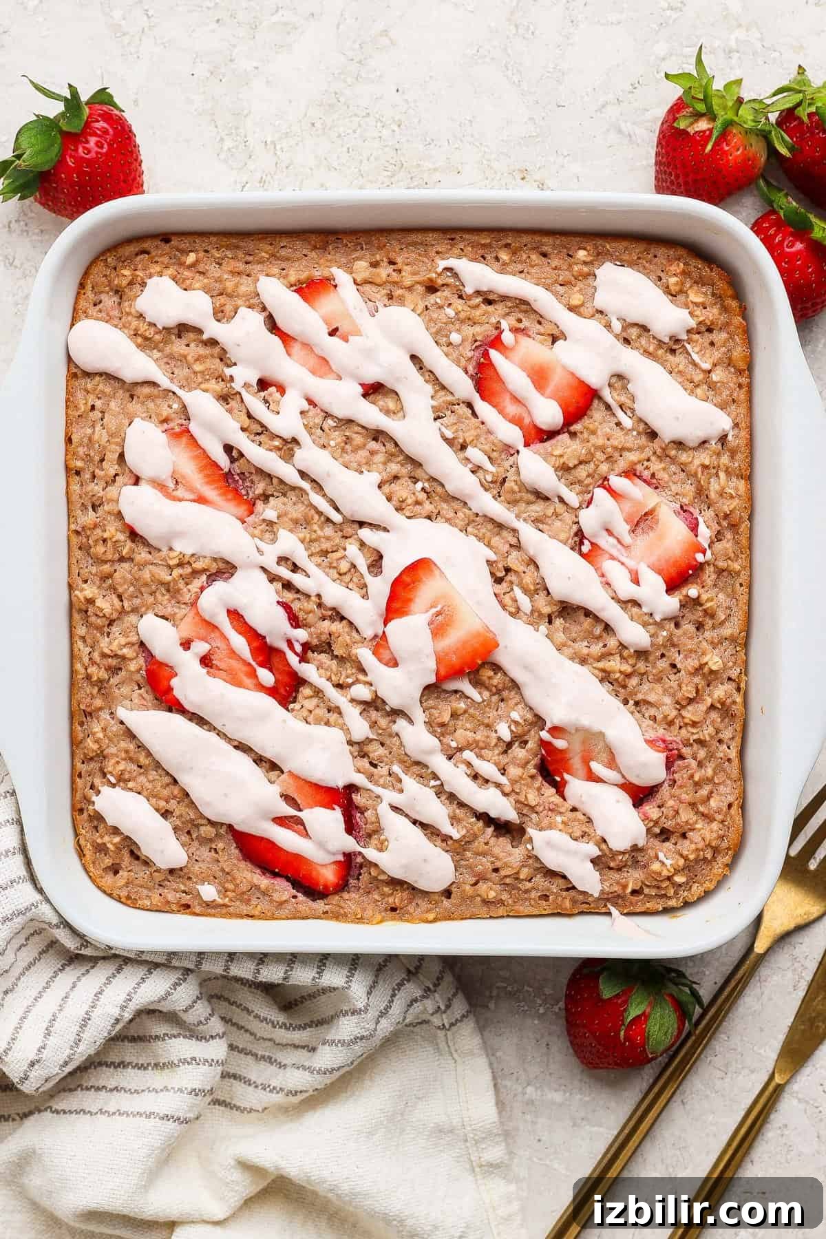 Strawberry Cheesecake Baked Oats Recipe with Creamy Filling 5 A square pan of baked oatmeal topped with sliced strawberries and white icing drizzle, set on a light surface with whole strawberries, a striped towel, and gold utensils nearby.