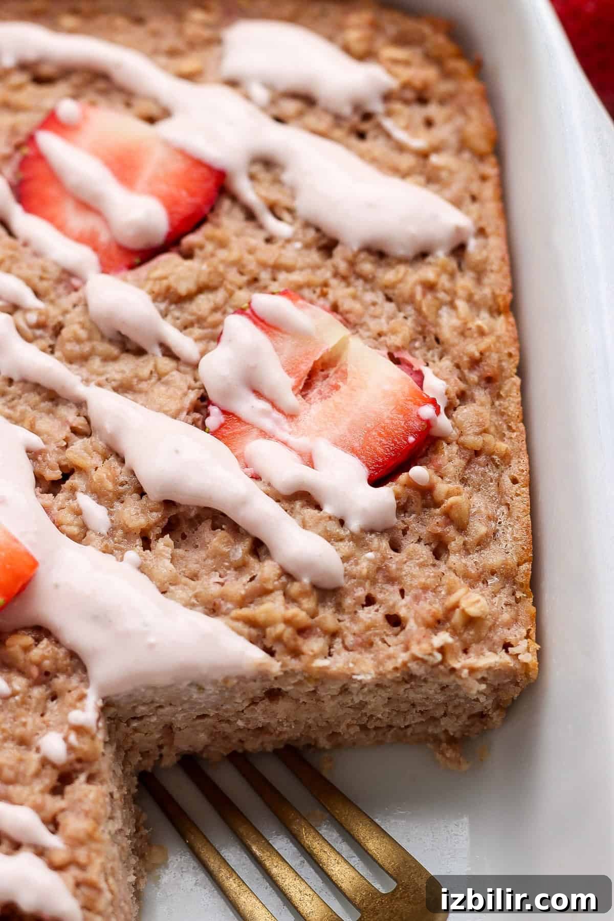 Strawberry Cheesecake Baked Oats Recipe with Creamy Filling 6 Baked oatmeal topped with sliced strawberries and white icing, shown in a baking dish with a serving removed and a metal spatula visible.