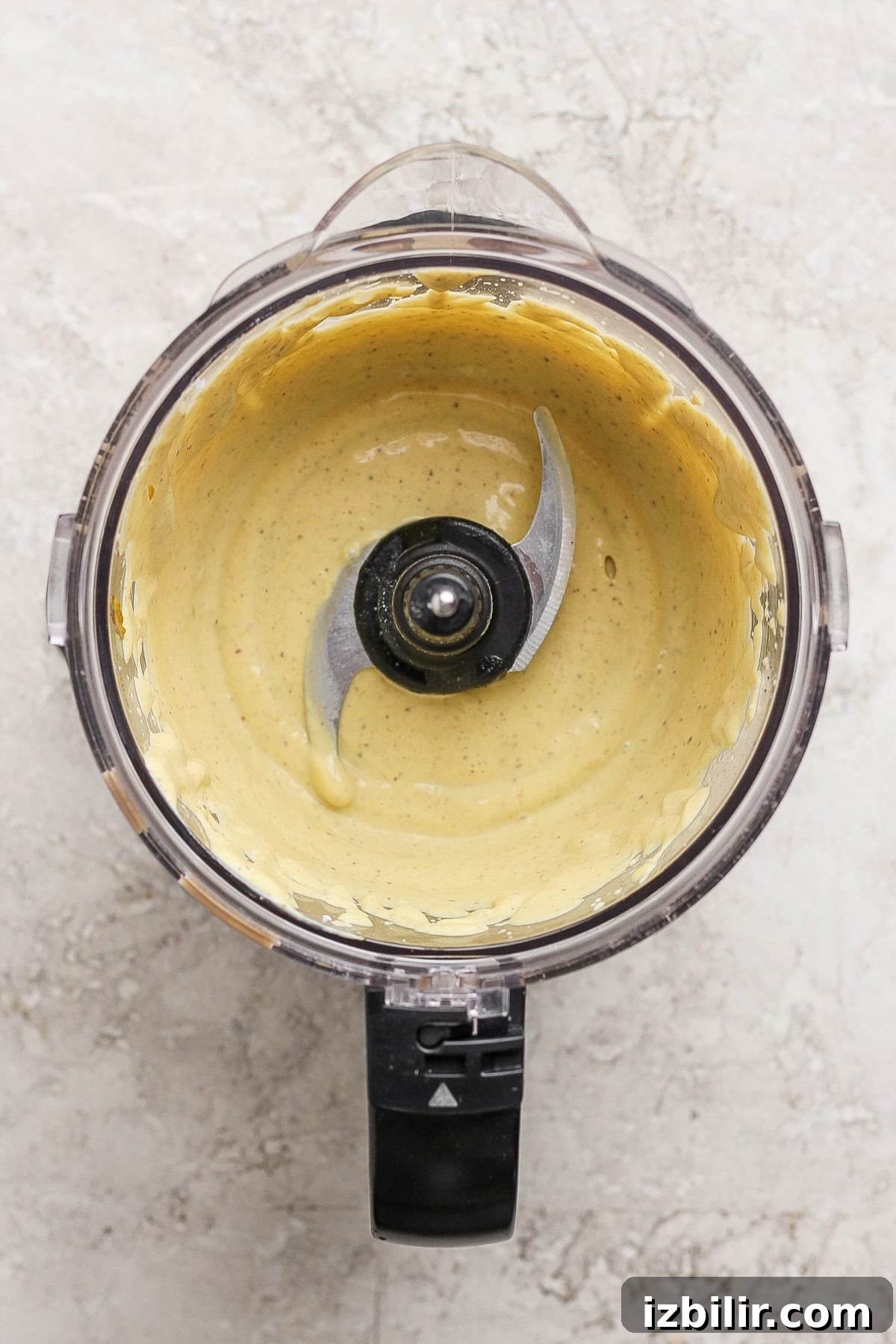 A food processor bowl containing a smooth, pale yellow creamy mixture with the blade visible in the center, set on a light textured surface.
