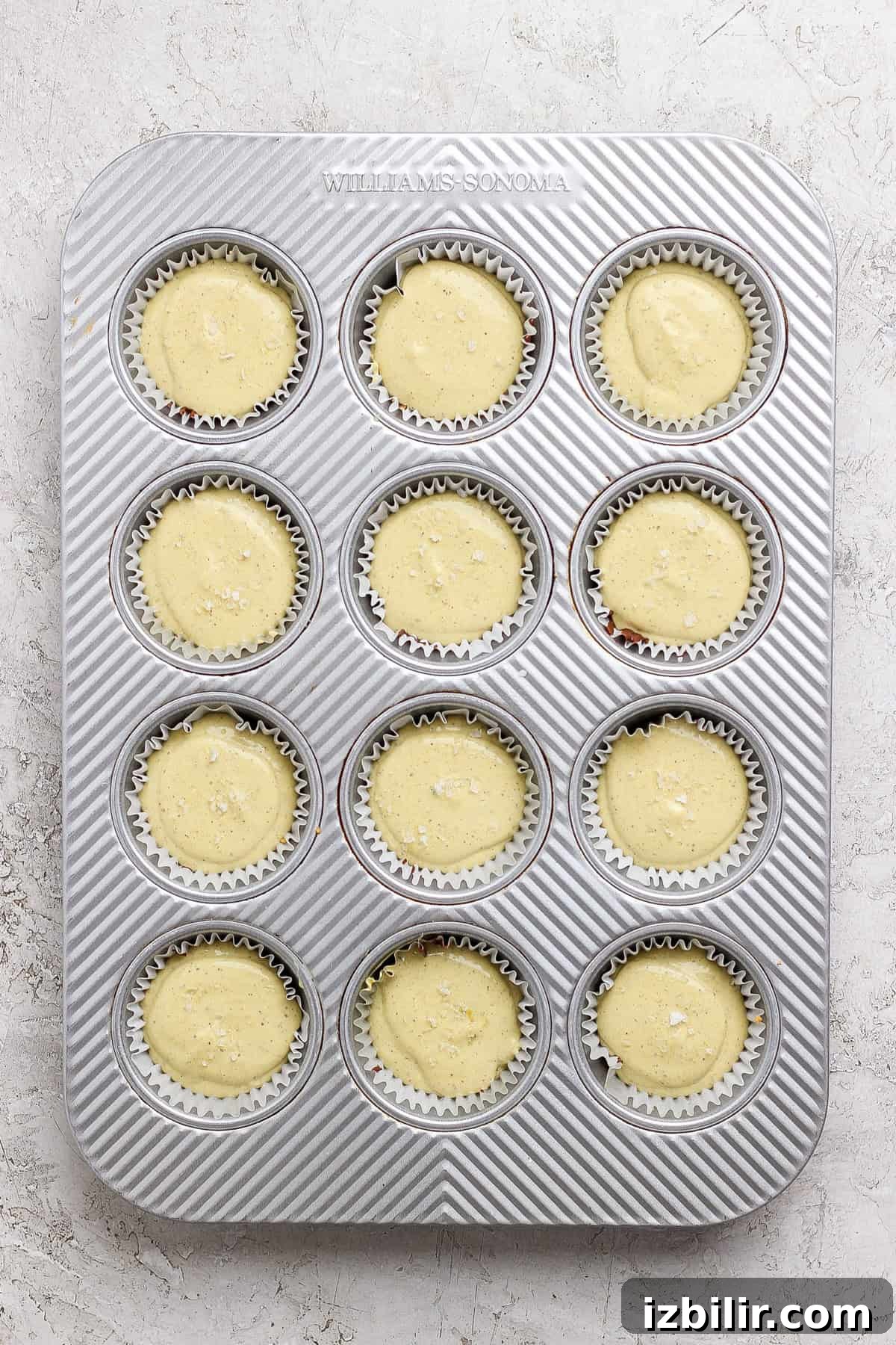 A metal muffin tin filled with twelve paper liners, each containing unbaked cupcake batter, sits on a light textured surface.