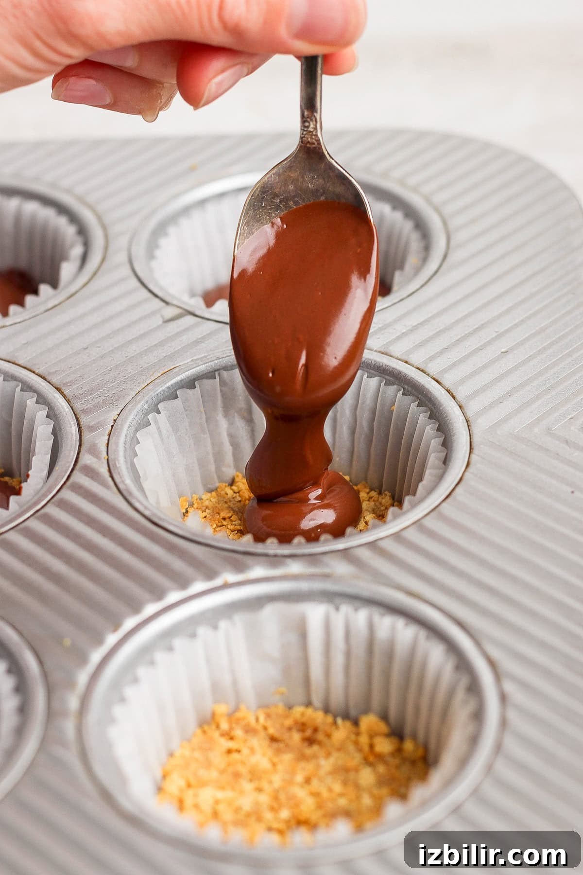 A hand holds a spoon pouring melted chocolate over crushed graham crackers in a paper-lined muffin tin.