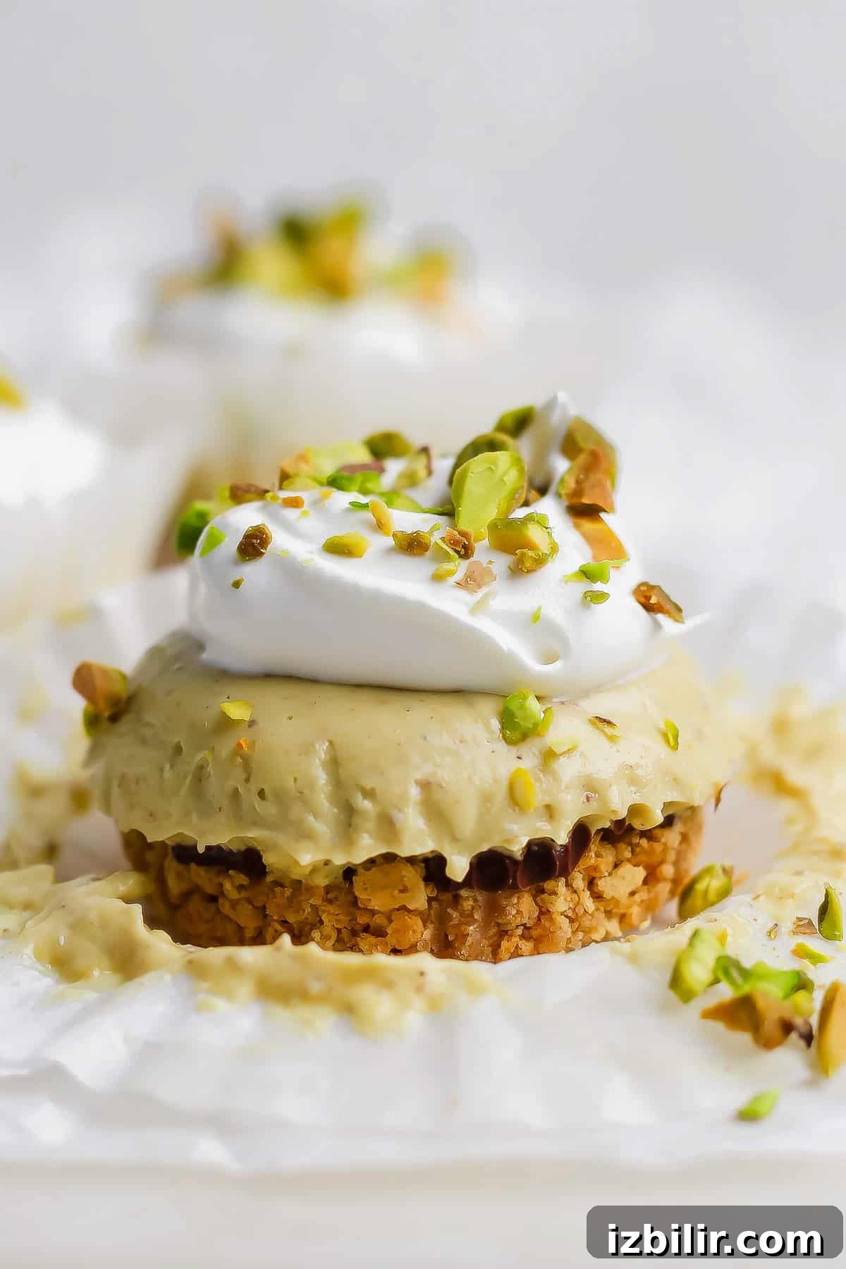 A mini cheesecake with a crumb crust, topped with whipped cream and chopped pistachios, sits on a piece of white parchment paper.