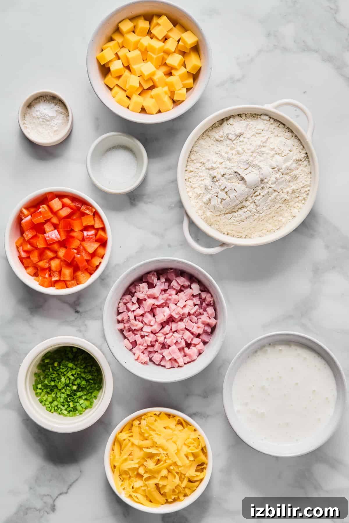 Assorted ingredients in bowls on a marble surface, including cubed cheddar cheese, flour, milk, diced ham, diced red bell pepper, grated cheese, chopped chives, salt, and baking powder.