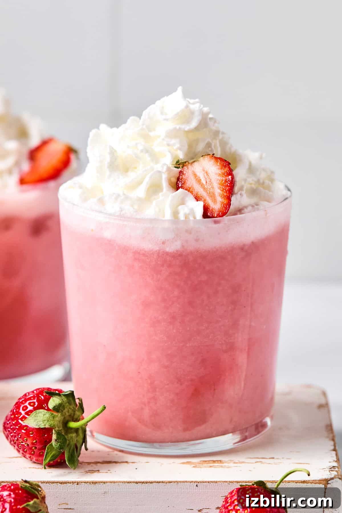 A glass of pink strawberry smoothie topped with whipped cream and a sliced strawberry, with whole strawberries nearby.