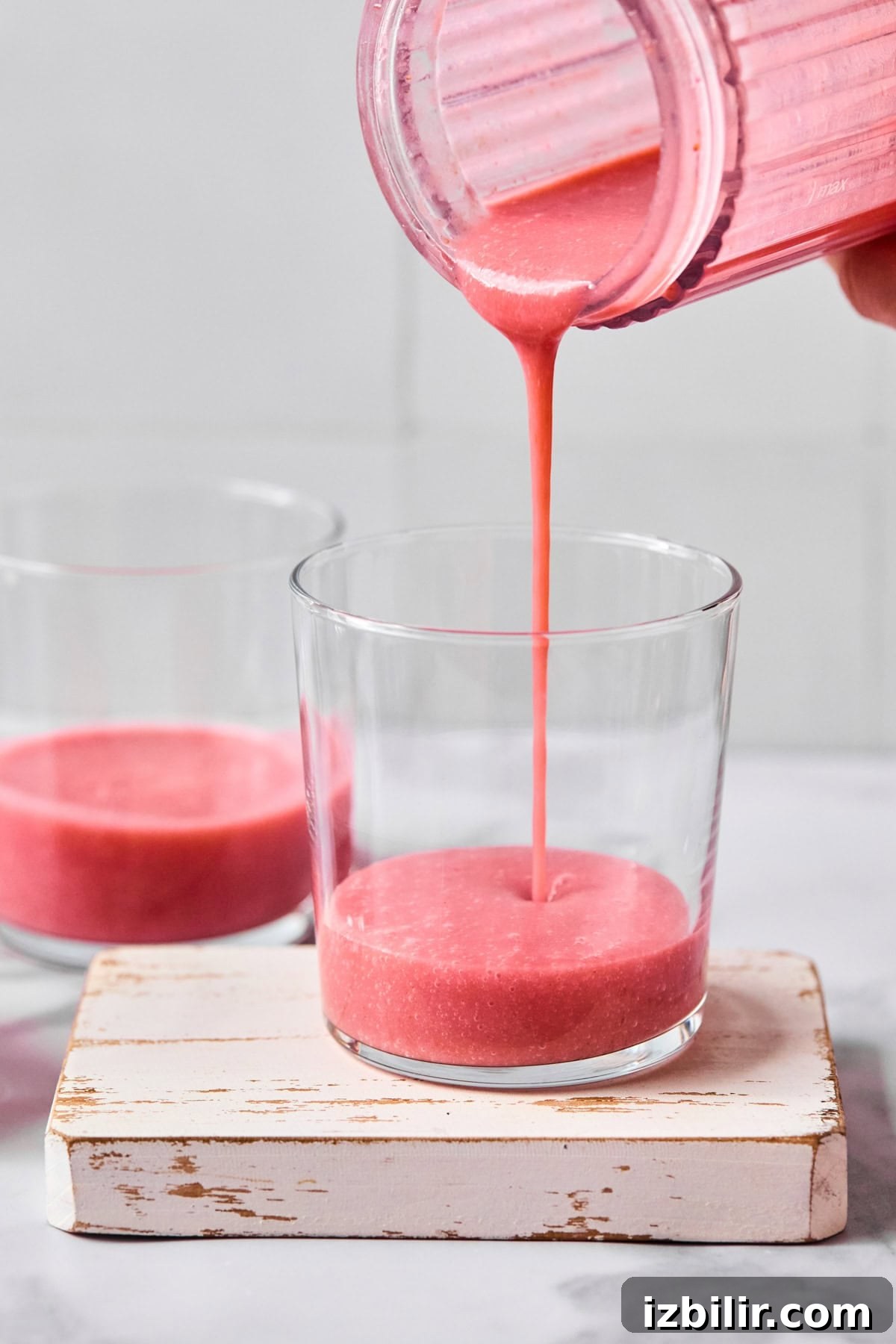 Pink smoothie being poured from a blender into a glass, with another glass of smoothie nearby on a small white wooden board.