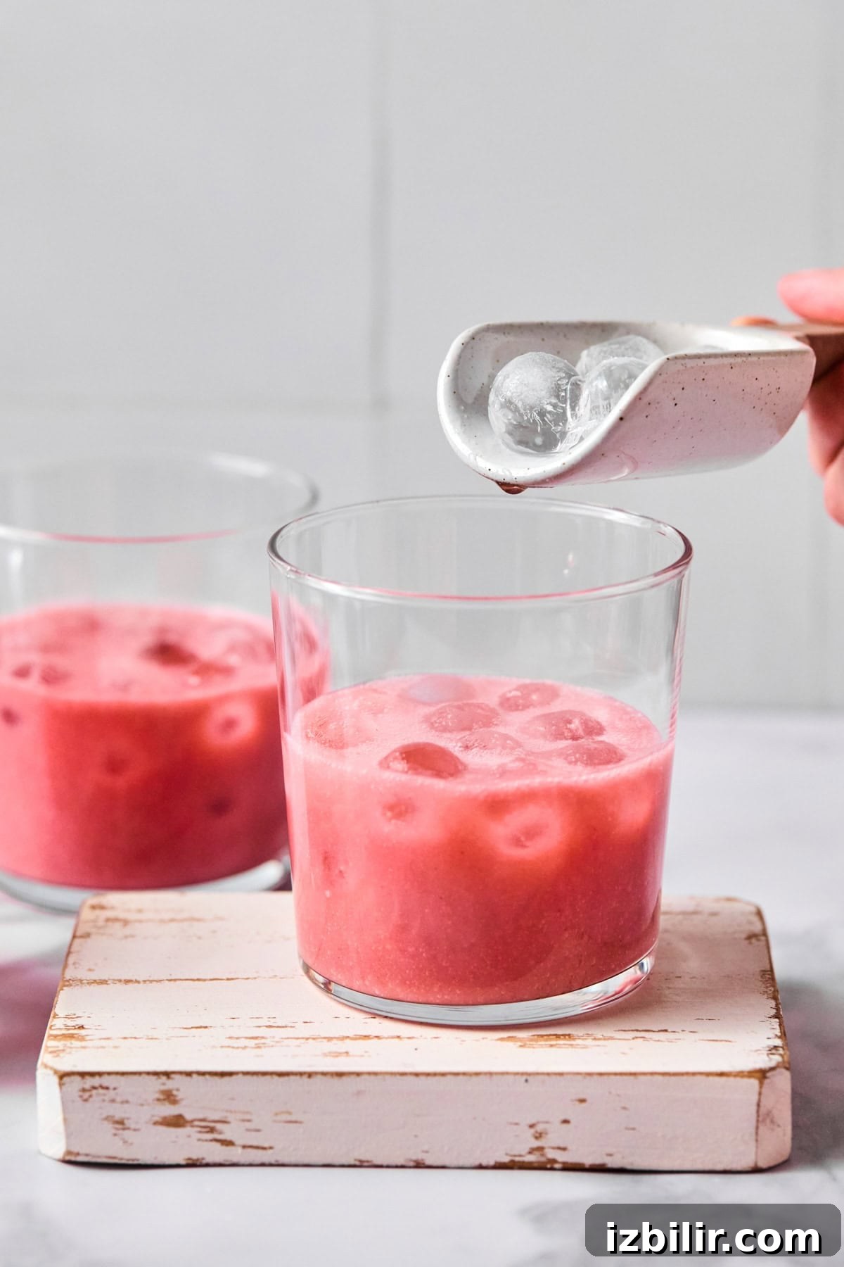 A hand adds ice cubes from a small white scoop into a glass of pink iced beverage, with another glass of the same drink in the background.