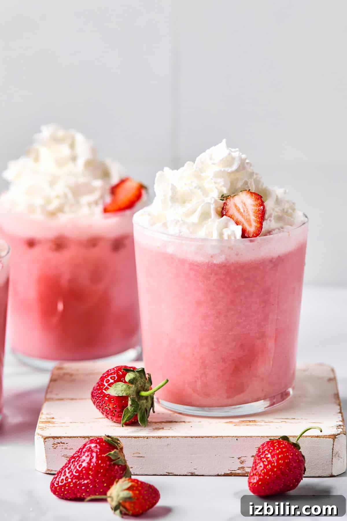 A glass of pink strawberry smoothie topped with whipped cream and a strawberry slice, with fresh strawberries placed beside the glass.