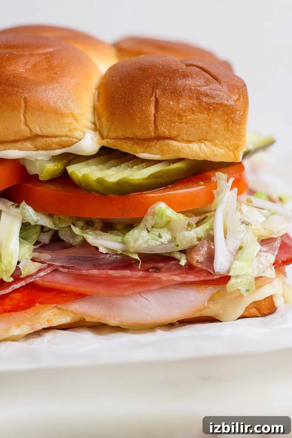 Italian Grinder Sliders Recipe: Classic Mini Italian Sandwiches 2 A close-up of a sandwich with sliced meats, shredded lettuce, tomato, pickles, mayonnaise, and a soft, browned bun.