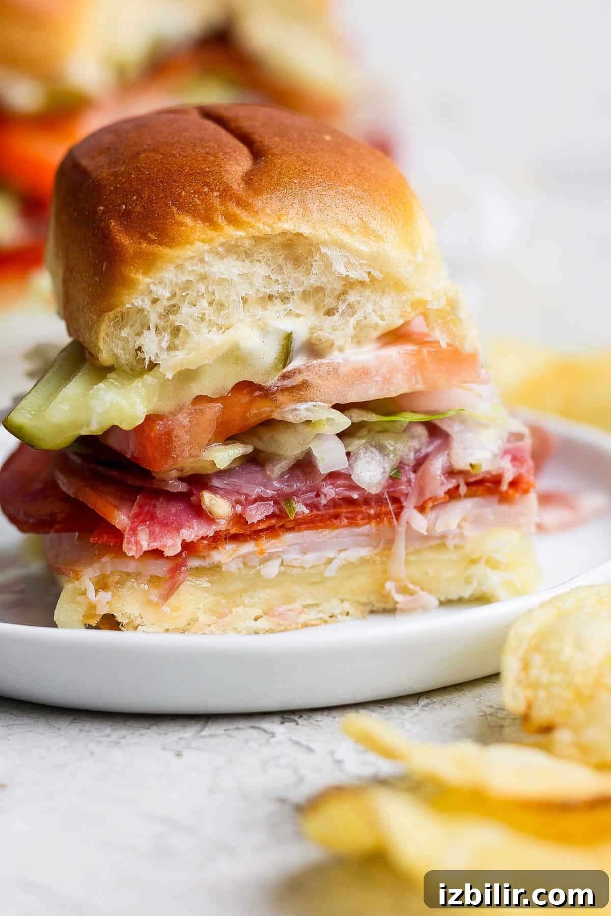 Italian Grinder Sliders Recipe: Classic Mini Italian Sandwiches 4 A small sandwich on a white plate, layered with deli meats, cheese, lettuce, tomato, and pickles, with a bite taken out. Potato chips are visible beside the plate.