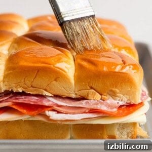 Italian Grinder Sliders Recipe: Classic Mini Italian Sandwiches 6 A hand brushes melted butter over the tops of stacked sandwich sliders with layers of deli meats and cheese on a baking tray.
