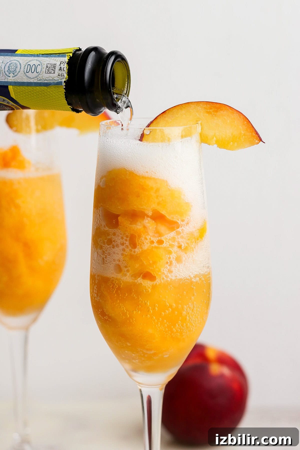 Frozen Peach Bellini: Sparkling Summer Cocktail 2 A glass filled with peach sorbet is being topped with sparkling wine, garnished with a peach slice; another glass and a peach are in the background.