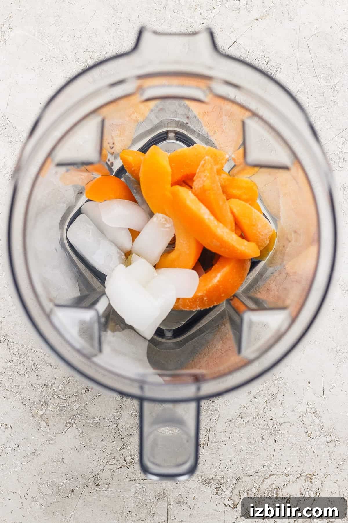 Frozen Peach Bellini: Sparkling Summer Cocktail 3 Top-down view of a blender containing ice cubes and orange fruit slices on a light-colored surface.