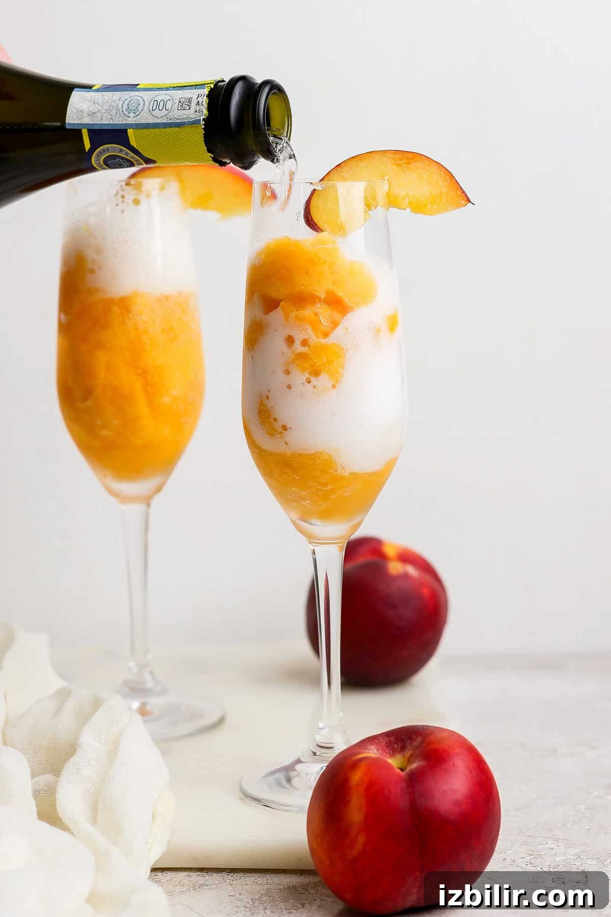 Frozen Peach Bellini: Sparkling Summer Cocktail 6 A glass of peach sorbet with sparkling wine being poured over it, garnished with a peach slice; another filled glass and whole peaches are in the background.