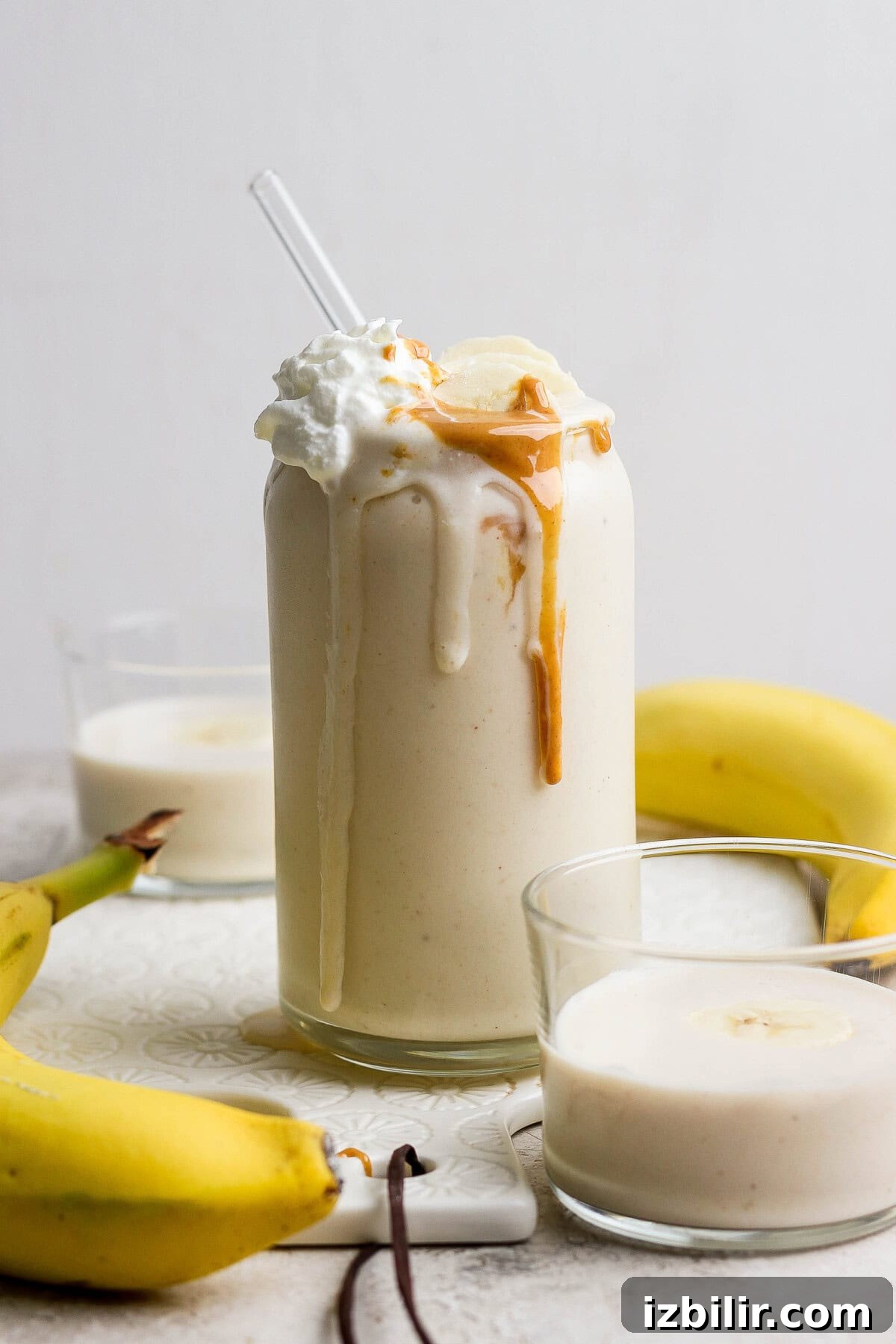Glass of banana smoothie topped with whipped cream and peanut butter, surrounded by bananas and two small glasses of smoothie.