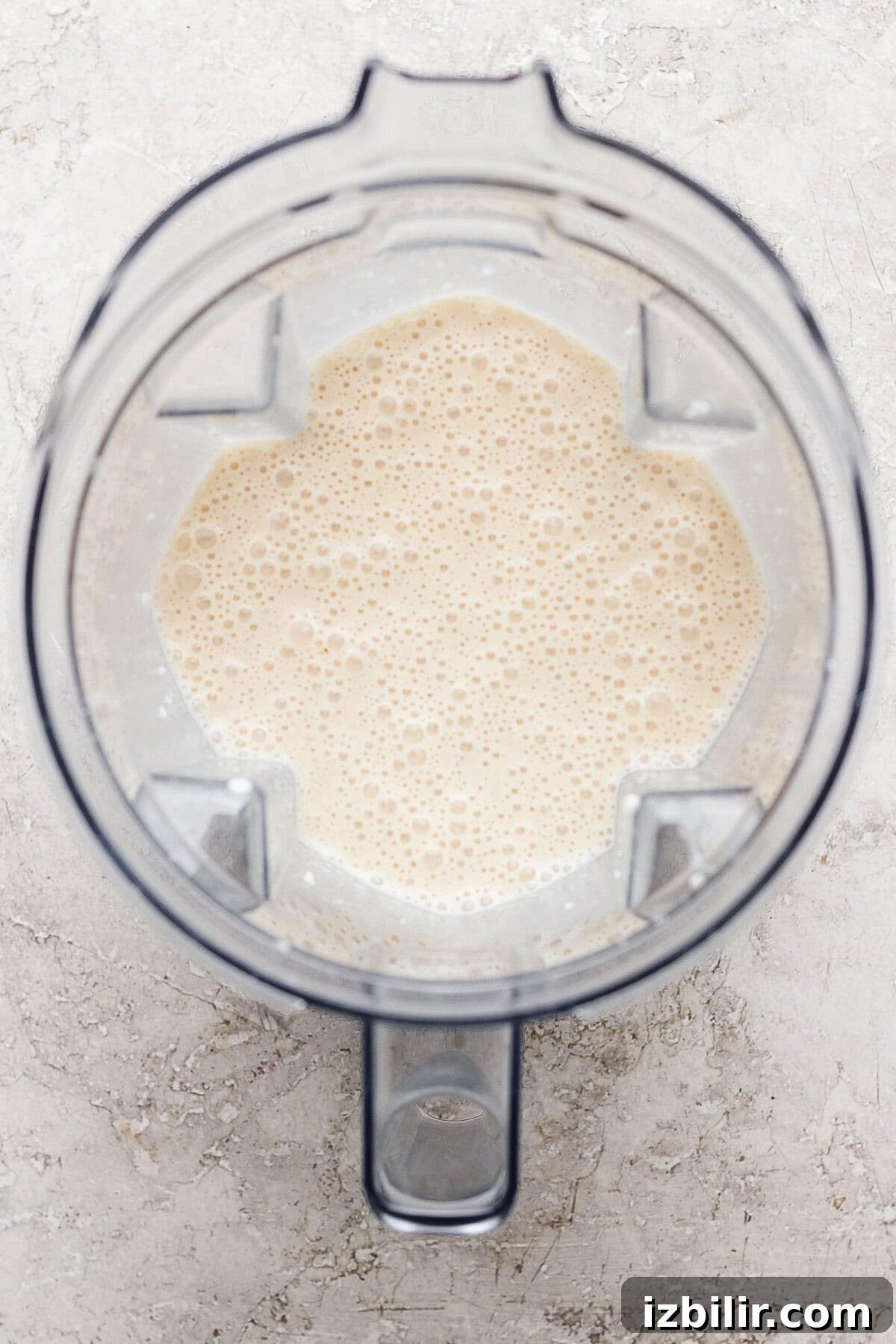 Top-down view of a blender containing a light beige, frothy liquid on a light textured surface.