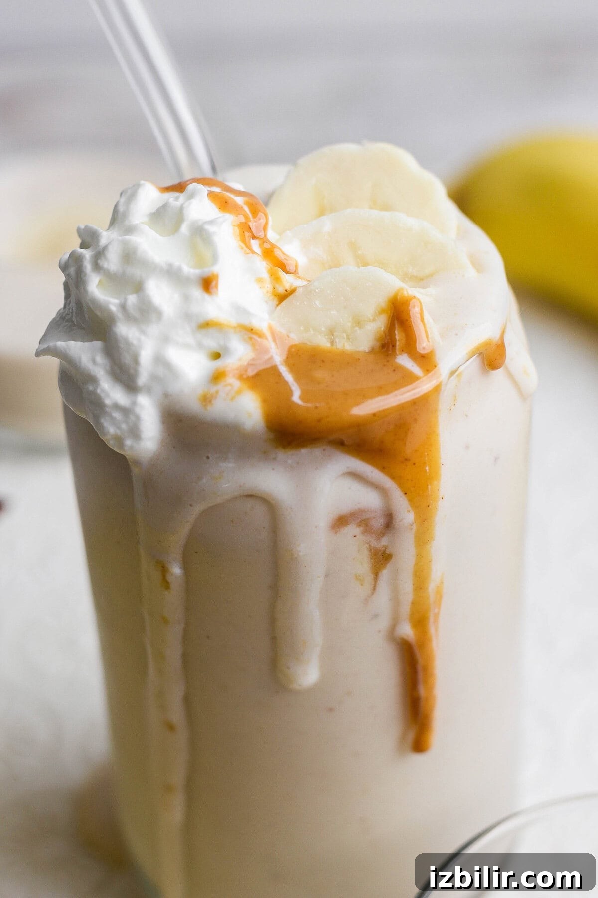 A glass of banana milkshake topped with whipped cream, banana slices, and peanut butter drizzle, with a straw inserted.