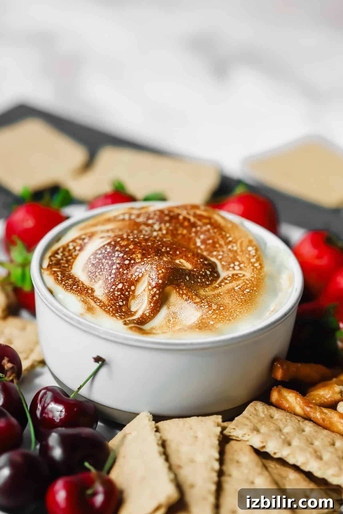 Homemade Maple Marshmallow Fluff Recipe 2 A white bowl filled with toasted marshmallow dip, surrounded by graham crackers, pretzels, strawberries, and cherries.