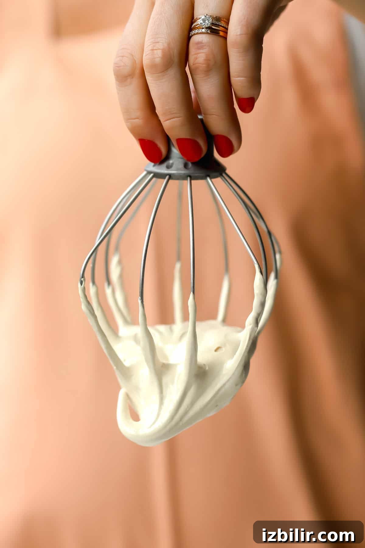Homemade Maple Marshmallow Fluff Recipe 4 A hand with red nail polish holds a metal whisk coated in thick, creamy batter against a peach-colored background.