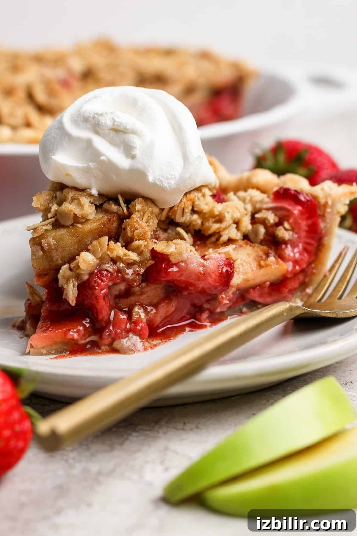 A slice of strawberry apple pie crumble topped with whipped cream sits on a white plate, with a gold fork, fresh strawberries, and apple slices alongside it.