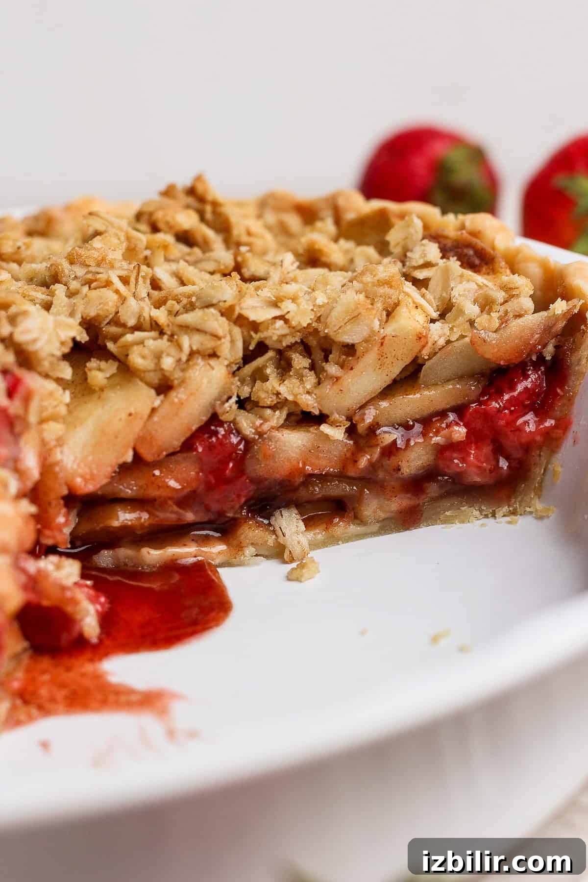 A close-up of a sliced strawberry apple pie crumble with a golden oat topping, filled with layers of apples and strawberries in a white dish.