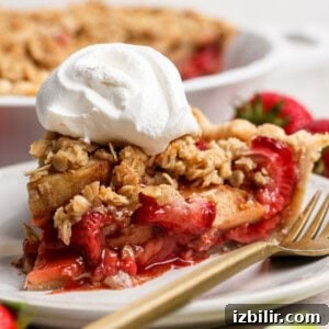 A slice of strawberry apple pie crumble topped with whipped cream sits on a white plate with a gold fork, fresh strawberries in the background.