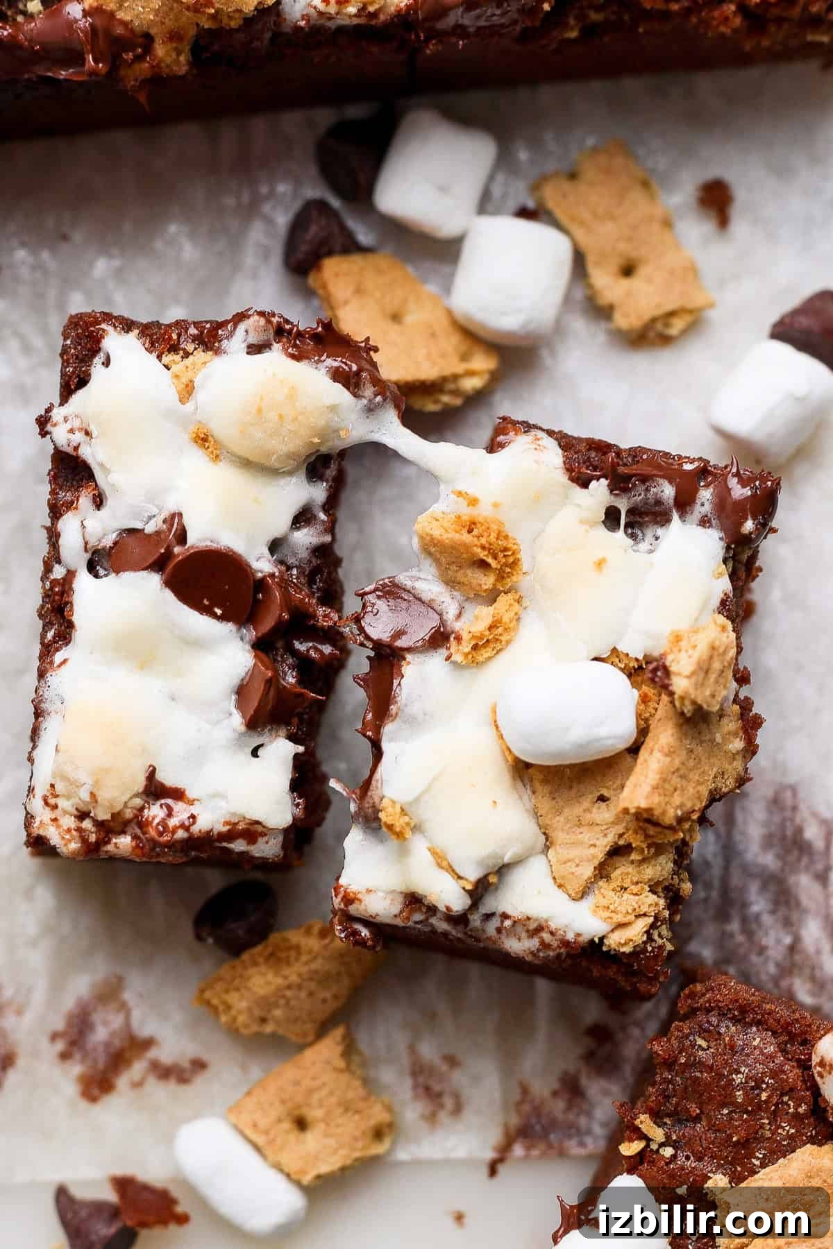 Protein S'mores Brownies Recipe 2 Two s'mores brownies topped with melted marshmallows, chocolate chips, and graham cracker pieces, placed on parchment paper with scattered ingredients around.