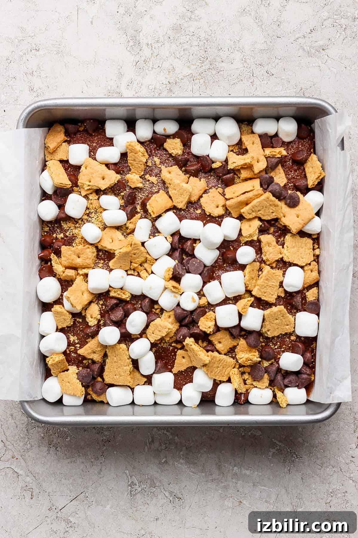 Protein S'mores Brownies Recipe 4 A square baking pan lined with parchment paper, filled with a mixture of mini marshmallows, chocolate chips, and broken graham crackers sits atop a rich s'mores brownies base.