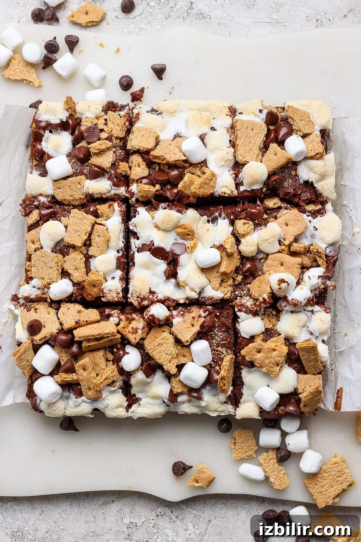 Protein S'mores Brownies Recipe 5 A batch of s'mores bars cut into squares, topped with mini marshmallows, graham cracker pieces, and chocolate chips on parchment paper—these treats are perfect for fans of classic s’mores brownies.