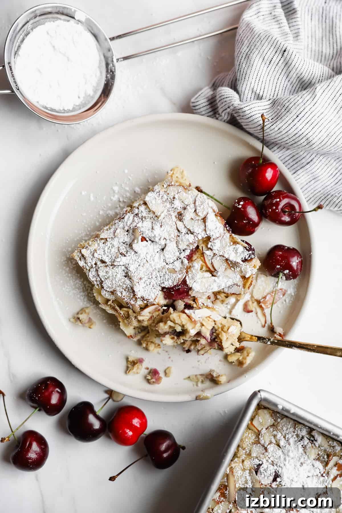 A slice of cherry dessert dusted with powdered sugar on a plate, surrounded by fresh cherries, a striped cloth, and a sifter with powdered sugar.