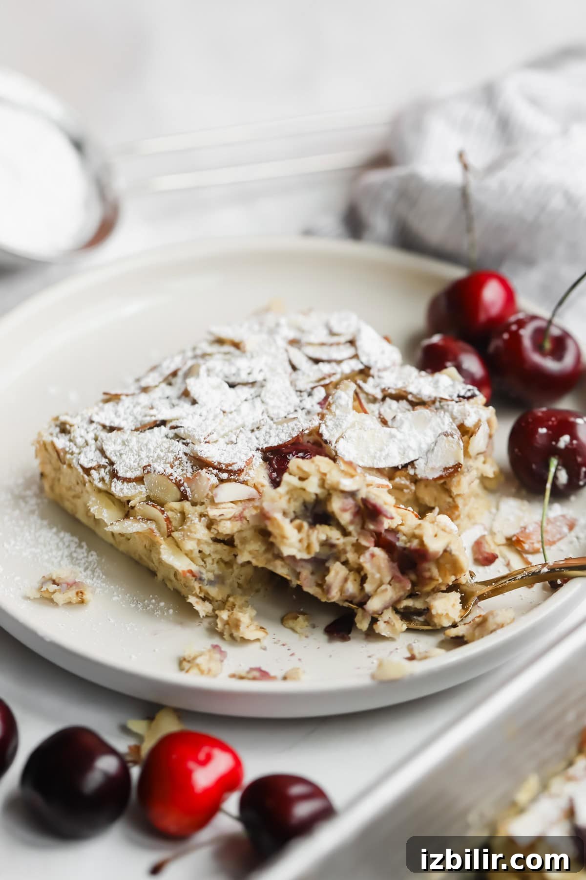 A partially eaten slice of almond and cherry cake topped with powdered sugar on a plate, with fresh cherries on the side.