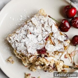 A square slice of almond cherry cake topped with powdered sugar on a plate, served with fresh cherries on the side.