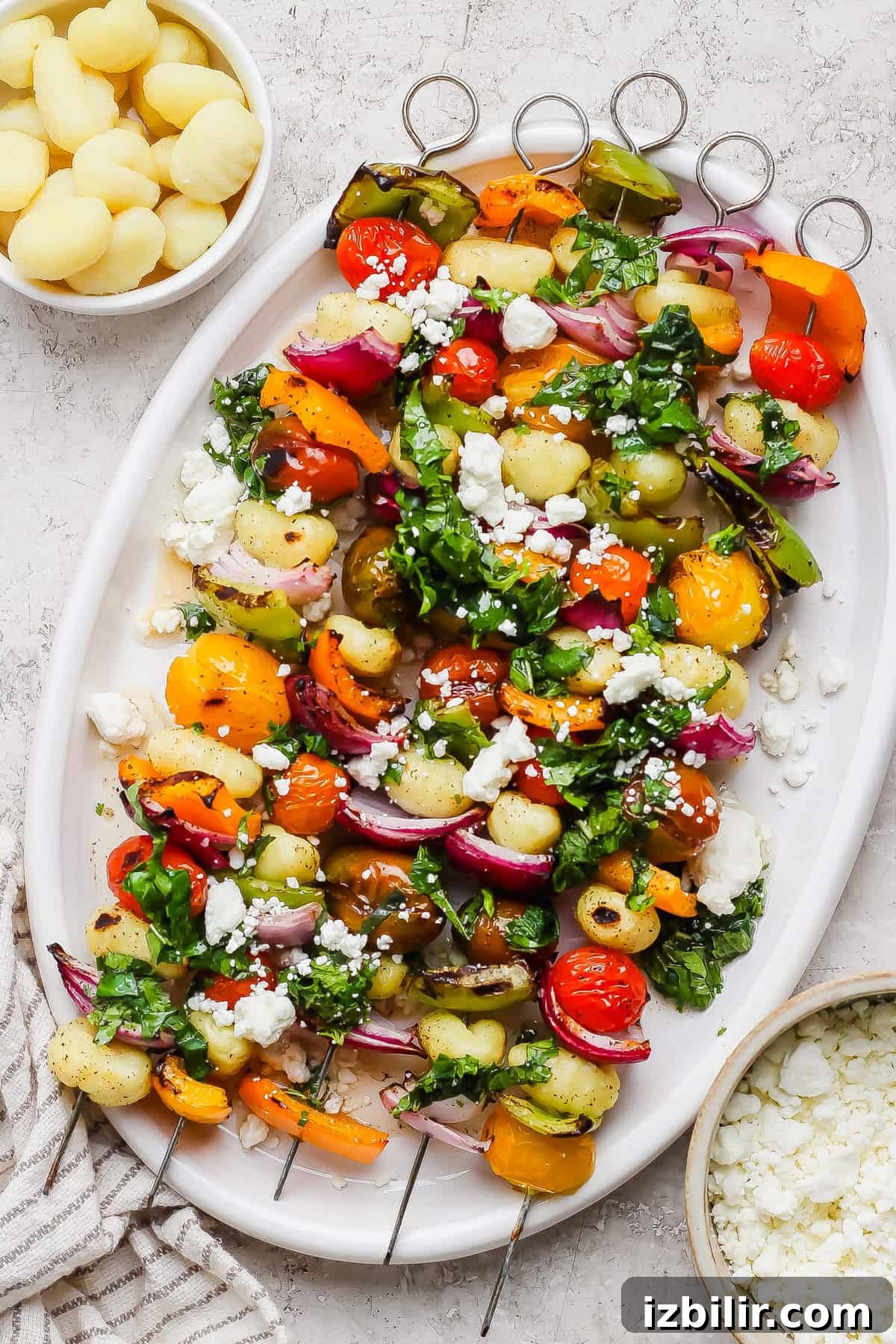 Rainbow Grilled Gnocchi Skewers with Roasted Veggies 2 A white platter with vegetable and gnocchi skewers, topped with chopped herbs and crumbled cheese, is pictured next to small bowls of cooked gnocchi and crumbled cheese.