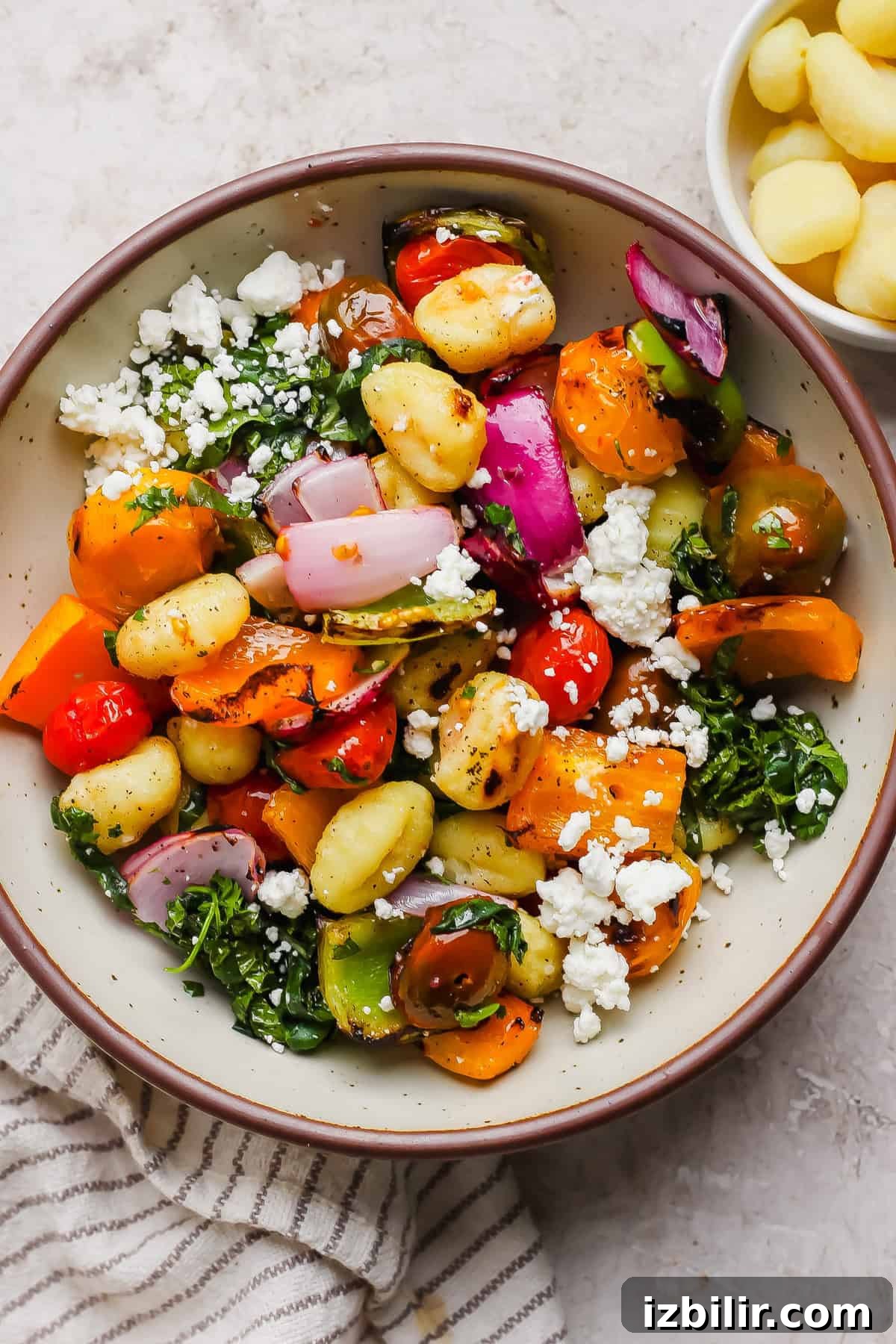 Rainbow Grilled Gnocchi Skewers with Roasted Veggies 4 A bowl of gnocchi mixed with roasted vegetables, red onion, tomatoes, leafy greens, and crumbled feta cheese, with a small bowl of gnocchi on the side.